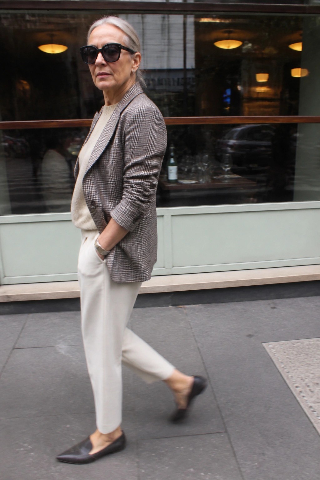 A woman over 60 in a brown and gray houndstooth blazer over a cream sweater with matching cream trousers and black pointed flats.