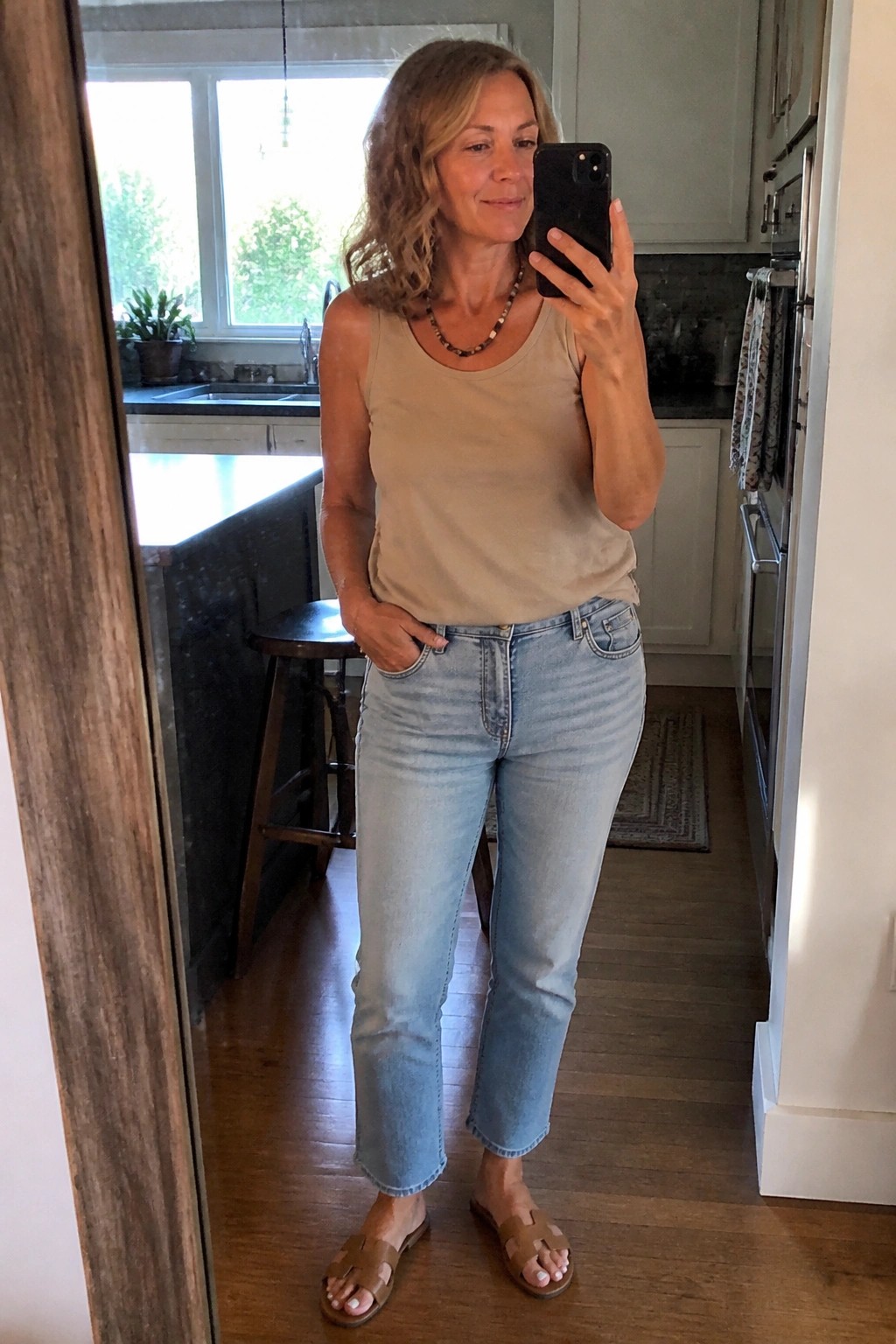 Woman in beige tank top and light blue jeans taking kitchen mirror selfie