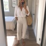 Woman in white linen shirt and pants holding phone and woven tote in bedroom mirror