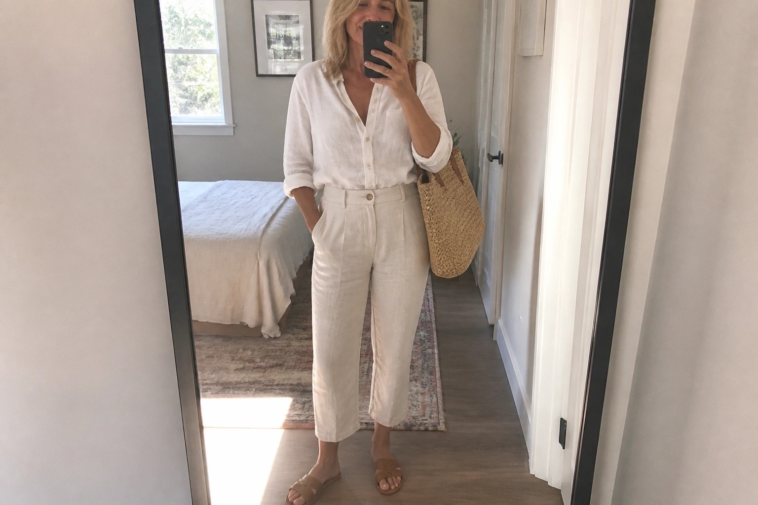 Woman in white linen shirt and pants holding phone and woven tote in bedroom mirror
