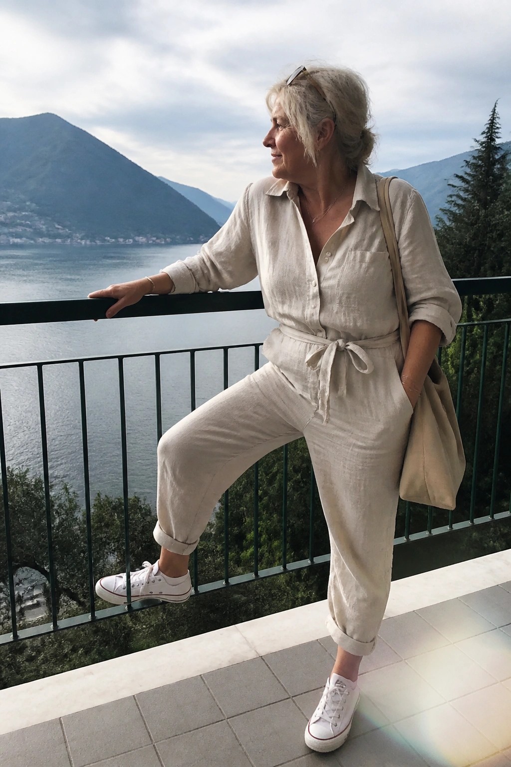 Gray-haired woman in beige jumpsuit posing on balcony overlooking mountain lake