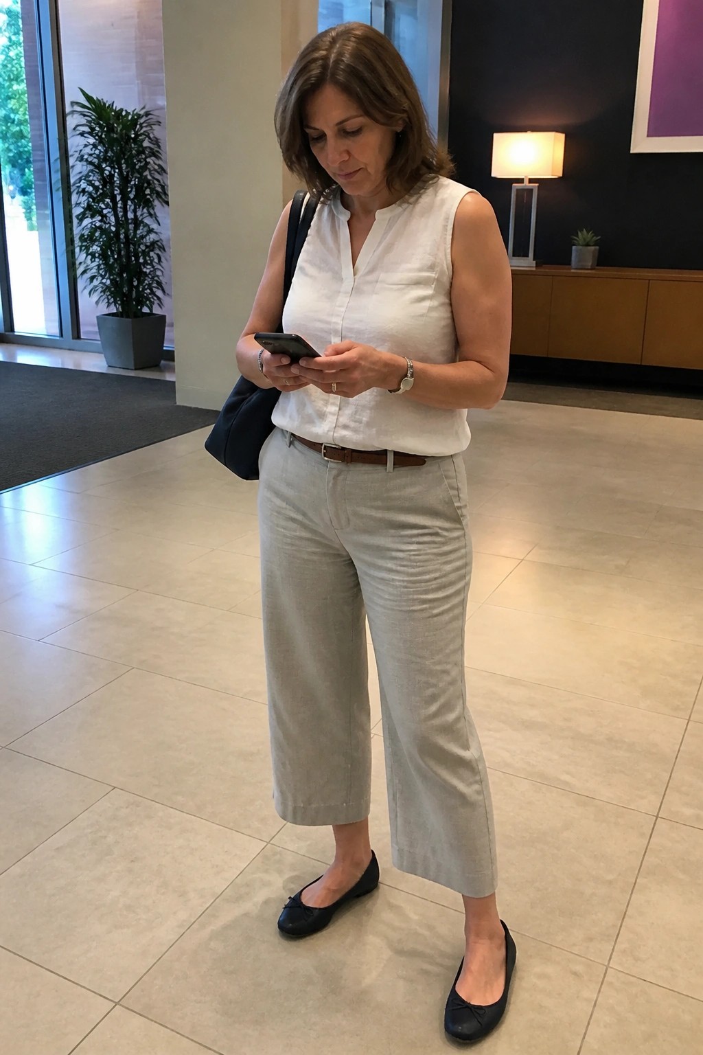 Woman in white blouse and gray pants checks smartphone in lobby