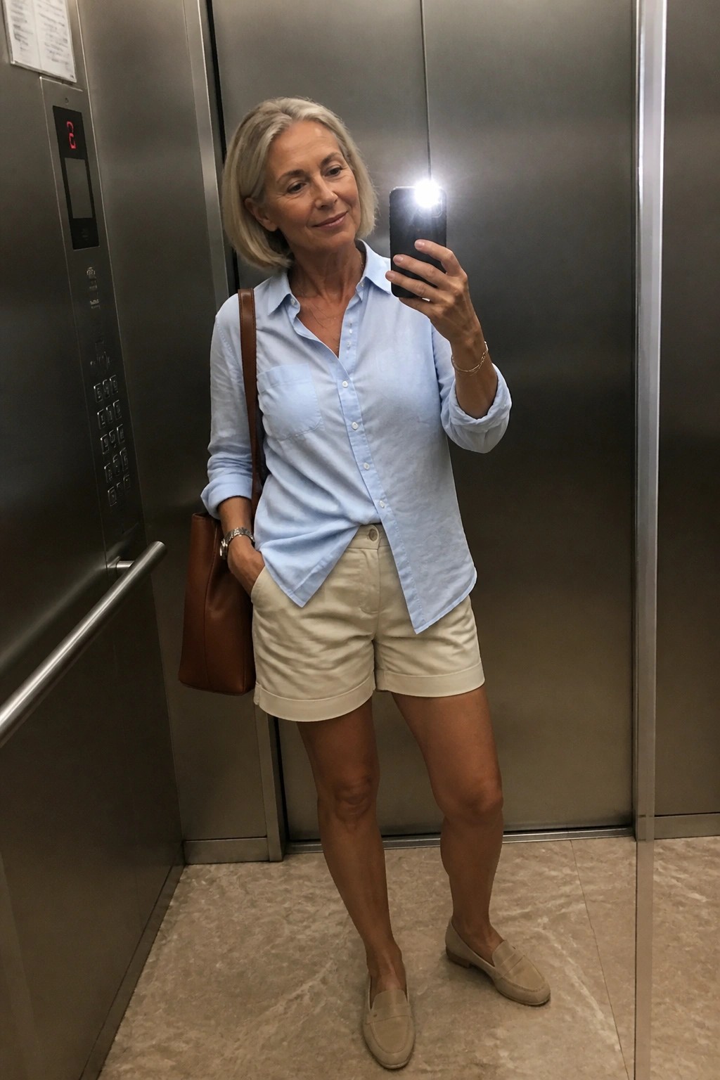Woman with gray hair taking elevator selfie in blue shirt and beige shorts.