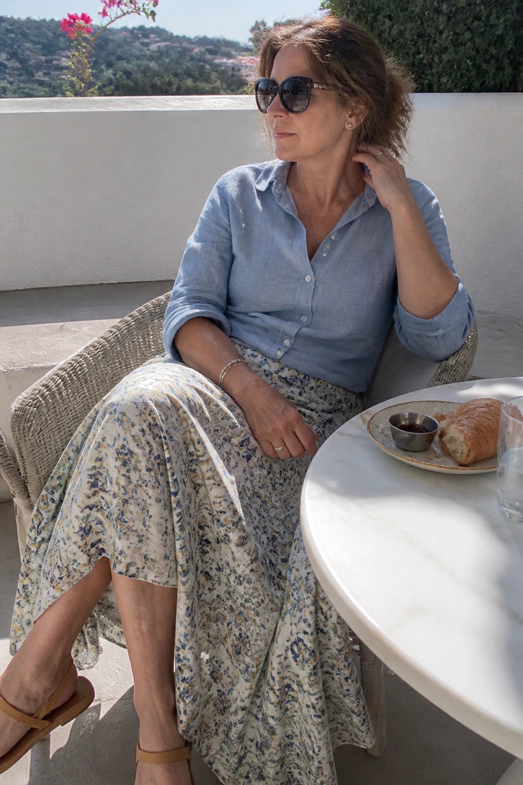 Woman in blue shirt and floral skirt seated outdoors with croissant and scenic view.
