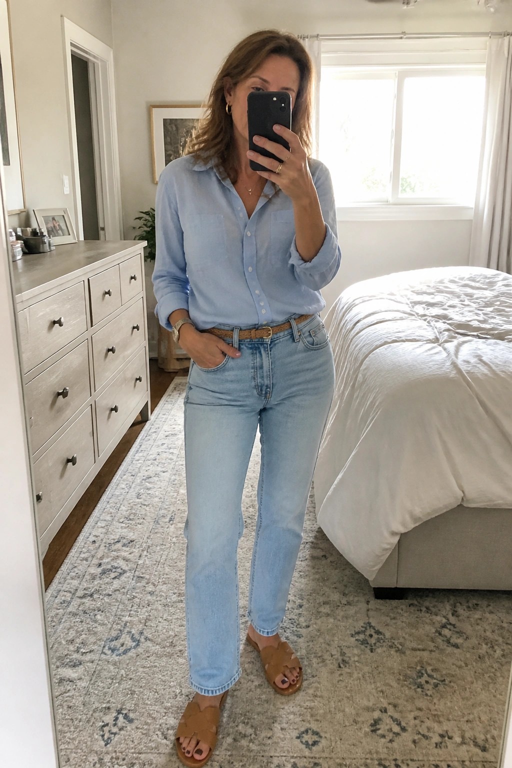 Woman wearing a light blue button-up shirt tucked into light blue jeans with a brown belt and brown slide sandals while taking a mirror selfie.