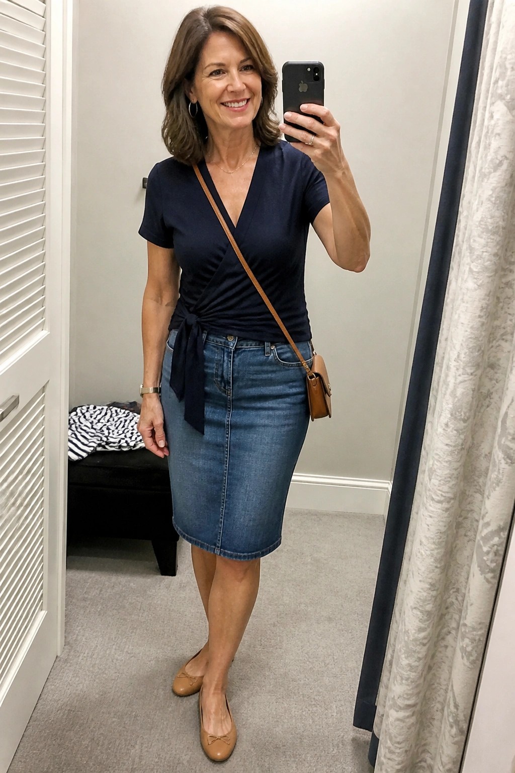 Woman in navy wrap top and denim skirt taking mirror selfie with phone