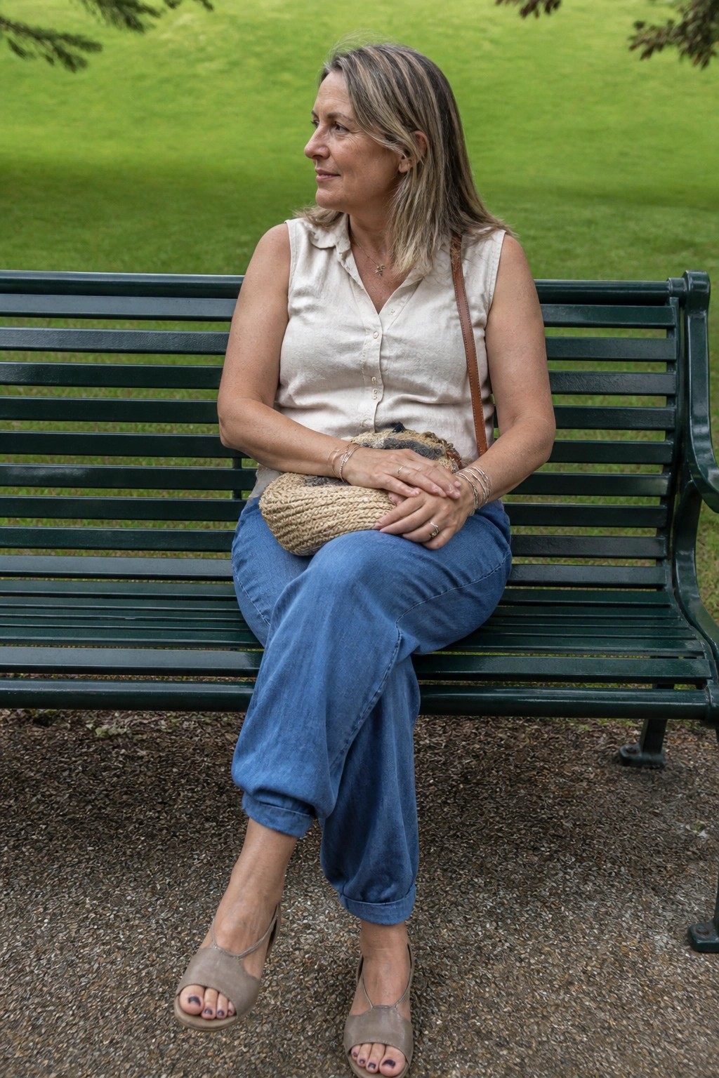 Woman over 50 in a sleeveless beige button-up top tucked into cuffed blue jeans with taupe sandals and a woven bag resting on her lap.