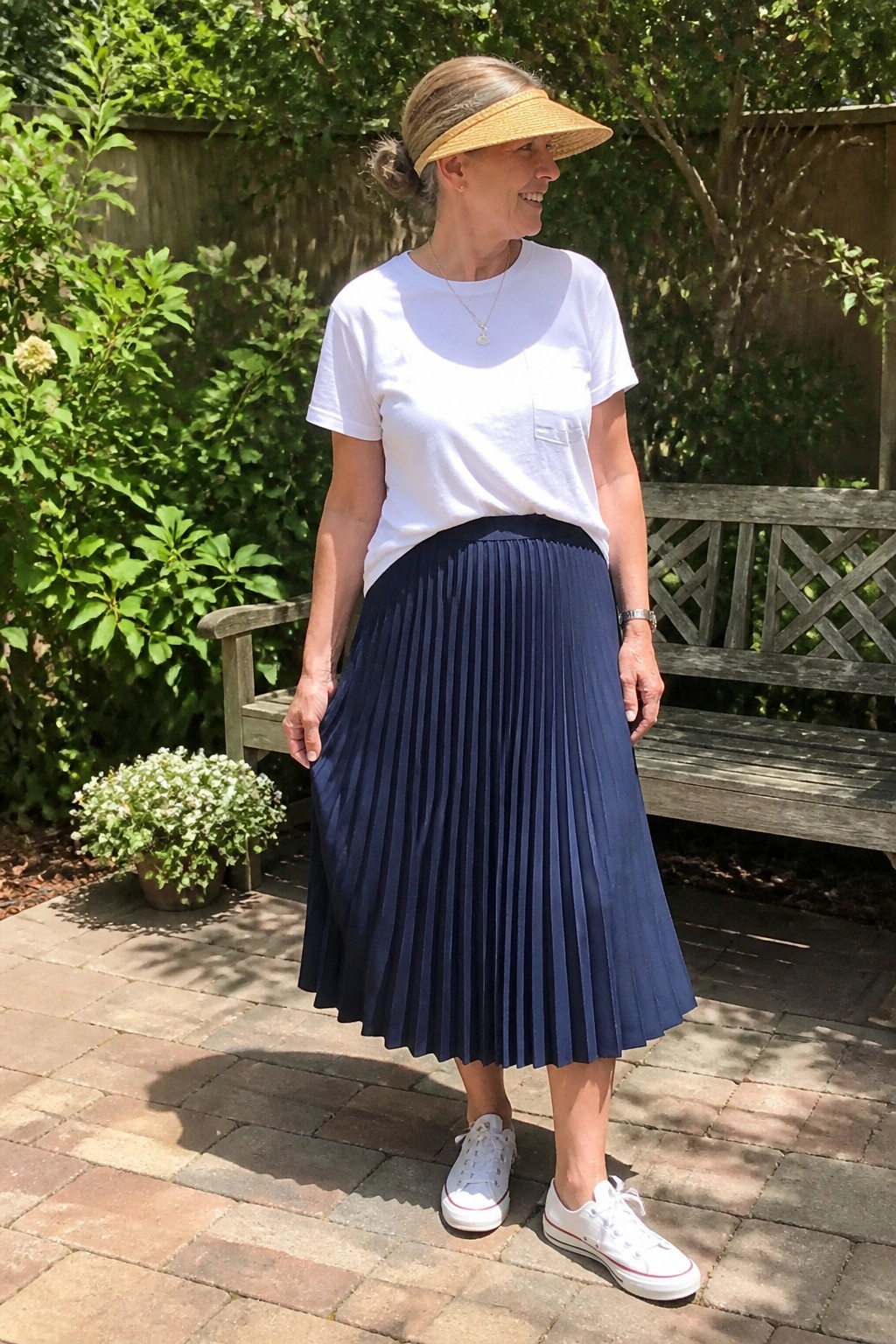 Woman over 50 in white short sleeve tee with pocket detail tucked into navy pleated midi skirt paired with white sneakers and straw visor standing on brick patio.