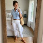 A woman over 50 in a light blue short sleeve button up shirt tucked into white cropped jeans with a woven brown belt, brown cross strap sandals, layered gold necklaces, and a large straw tote bag while taking a mirror selfie.