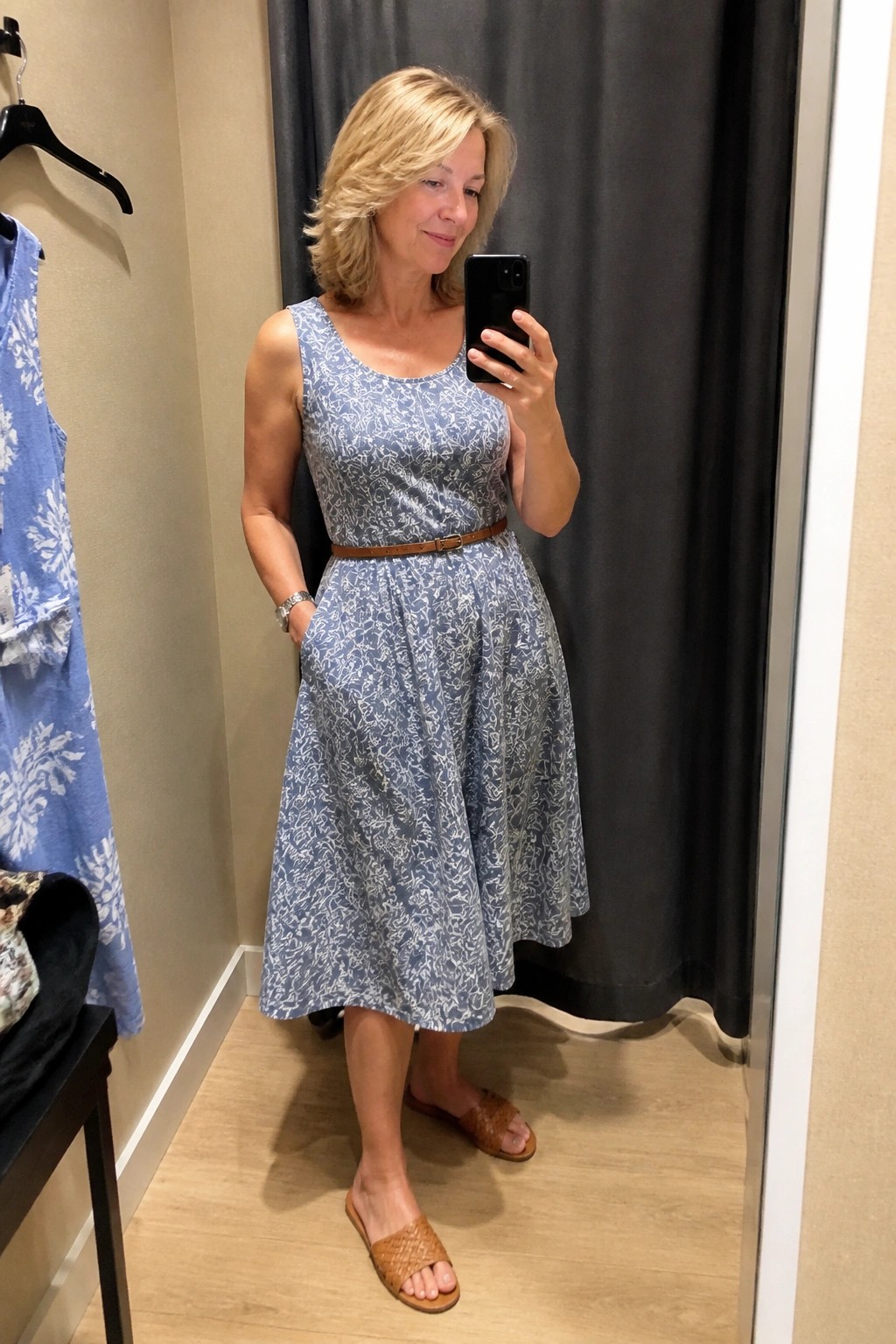 Woman over 50 wearing a sleeveless light blue and white patterned midi dress cinched with a thin brown belt and paired with brown woven slide sandals while taking a mirror selfie in a fitting room.