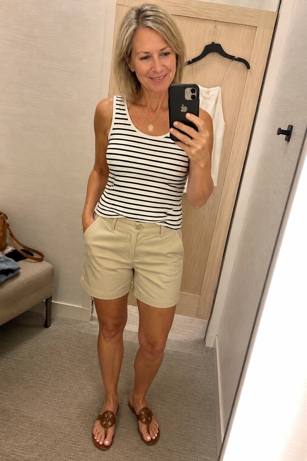 Woman in striped tank top and khaki shorts taking mirror selfie in fitting room