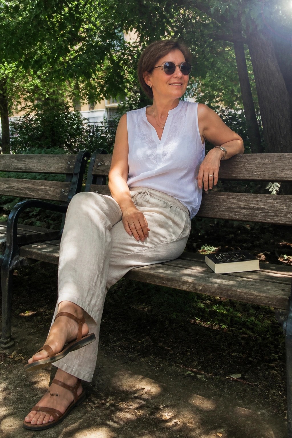 Woman in white top and beige pants relaxes on park bench with book
