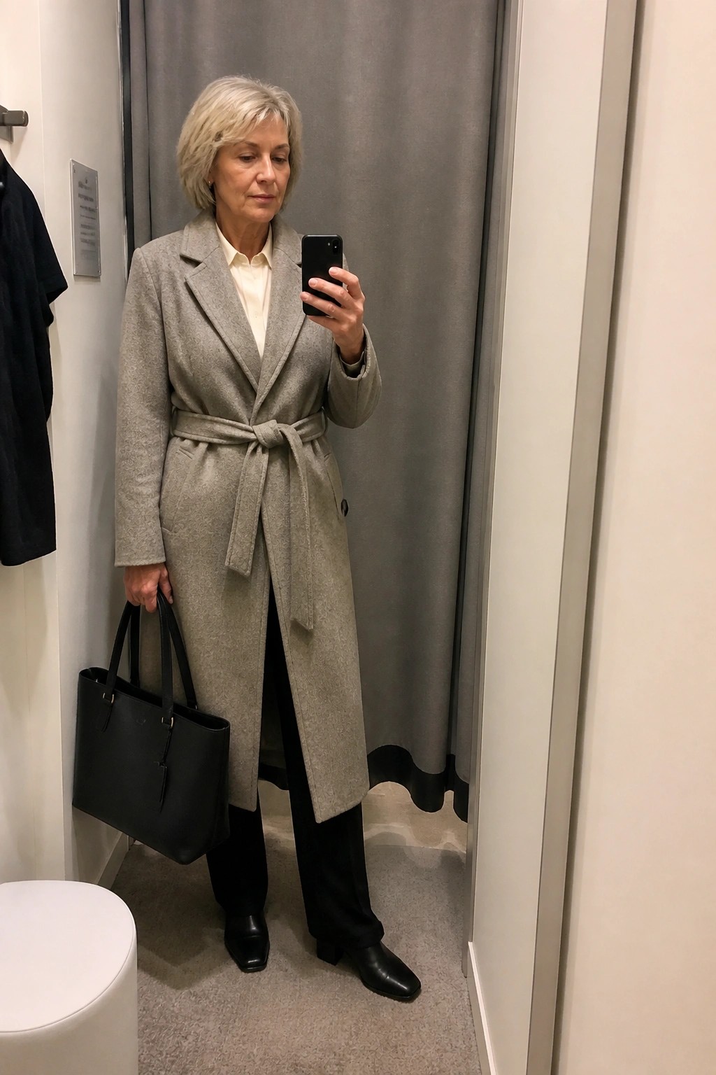 Woman in gray belted coat holding phone and black bag in fitting room.