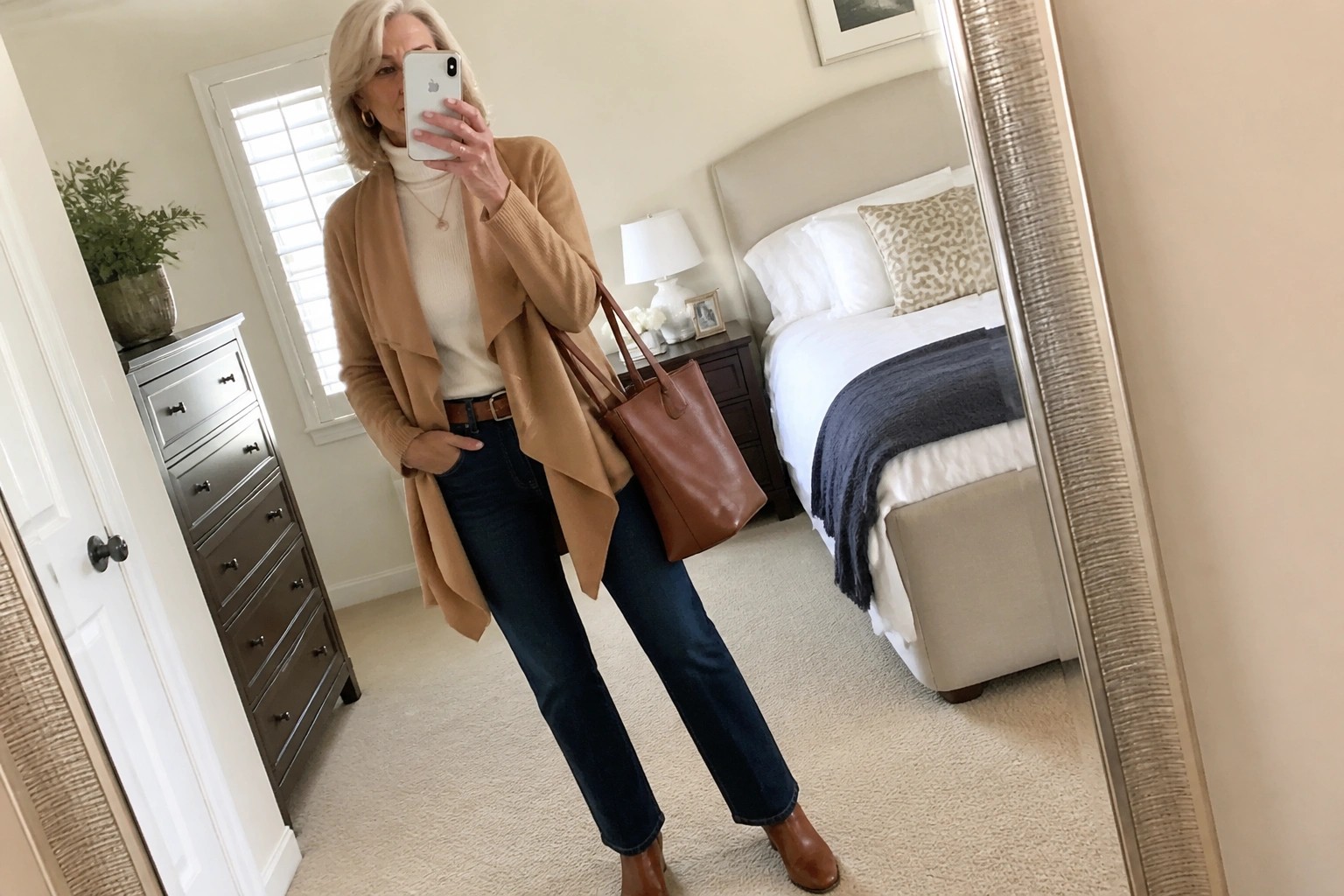 Silver-haired woman in cream turtleneck, camel cardigan, and jeans holding phone and bag