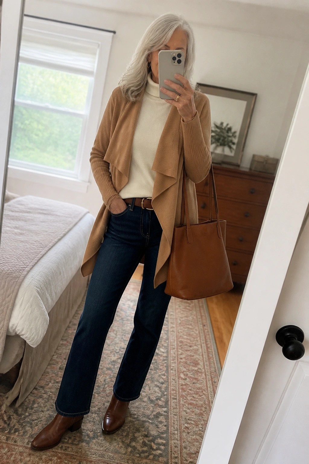 Silver-haired woman in cream turtleneck, camel cardigan, and jeans holding phone and bag