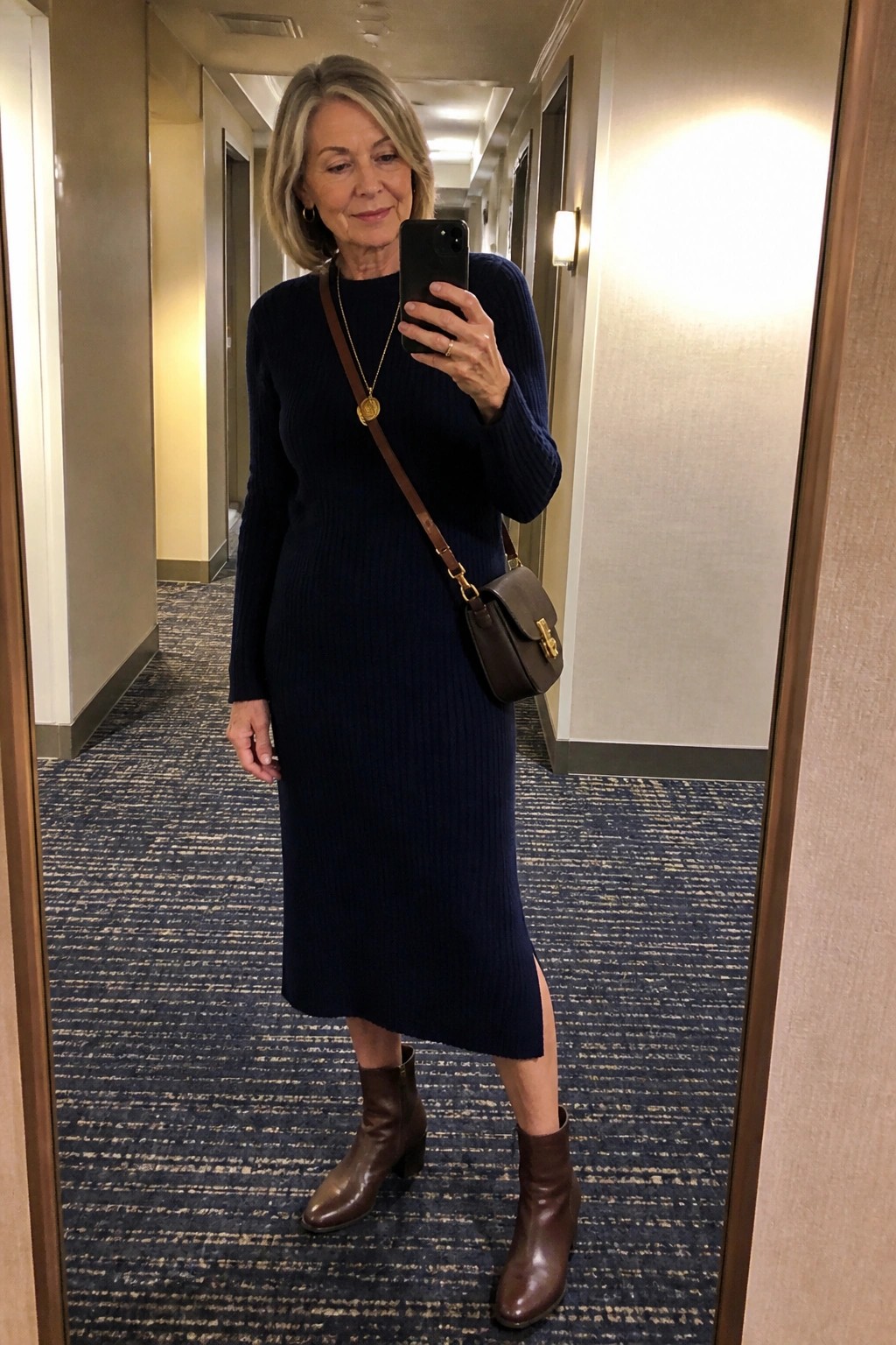 Woman wearing navy ribbed midi sweater dress with brown ankle boots and matching crossbody bag while holding phone for mirror selfie
