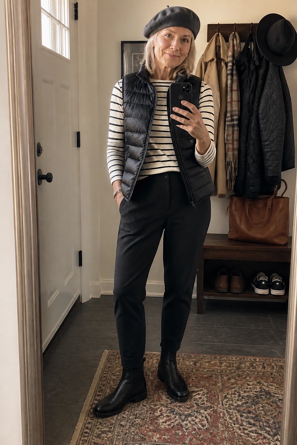 Woman in black beret and puffer vest taking mirror selfie in entryway with coats