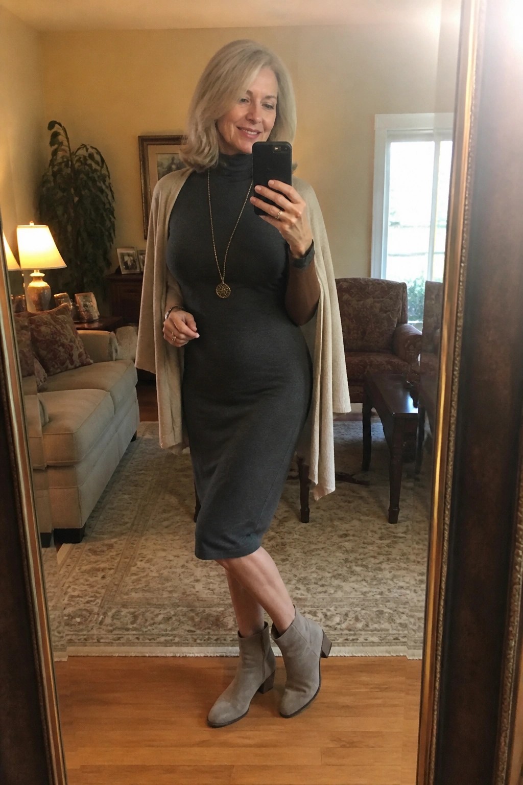 Woman in gray dress and beige cardigan taking mirror selfie in living room