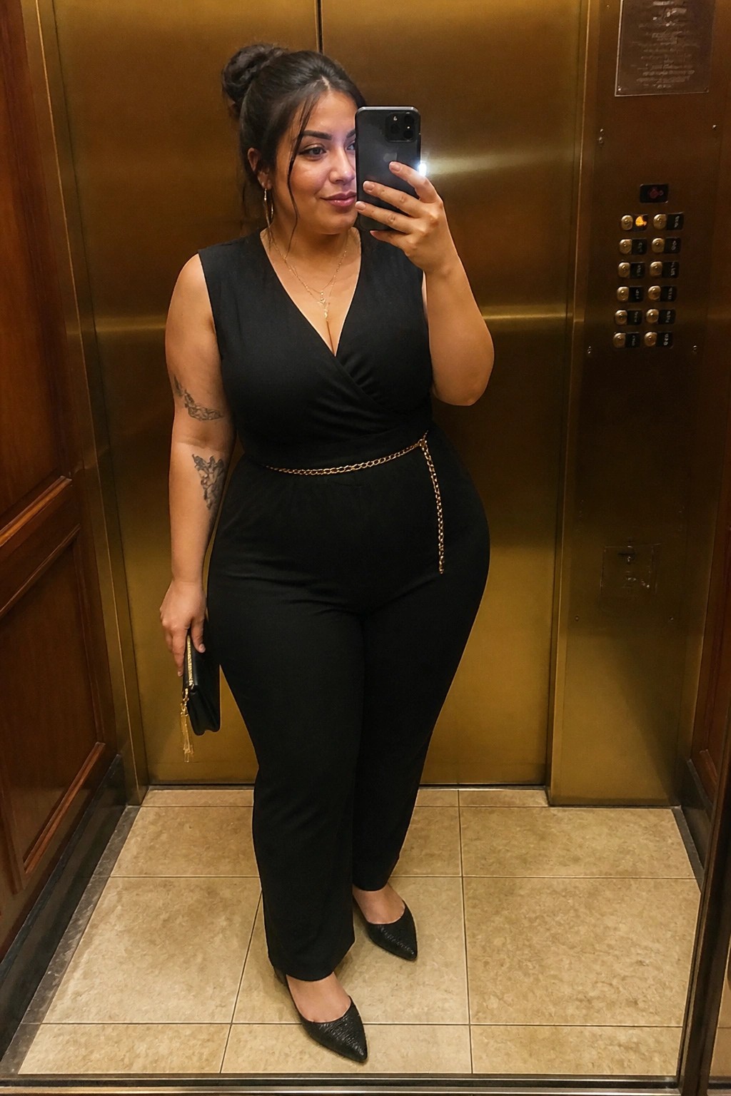 Woman in black jumpsuit with gold chain belt taking elevator selfie