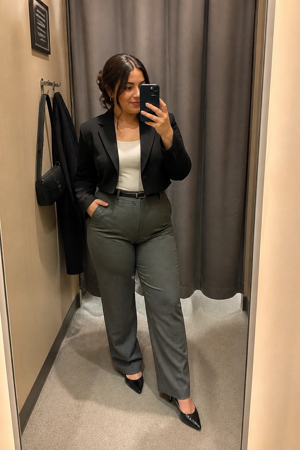 Woman in black blazer and gray trousers taking mirror selfie in fitting room.