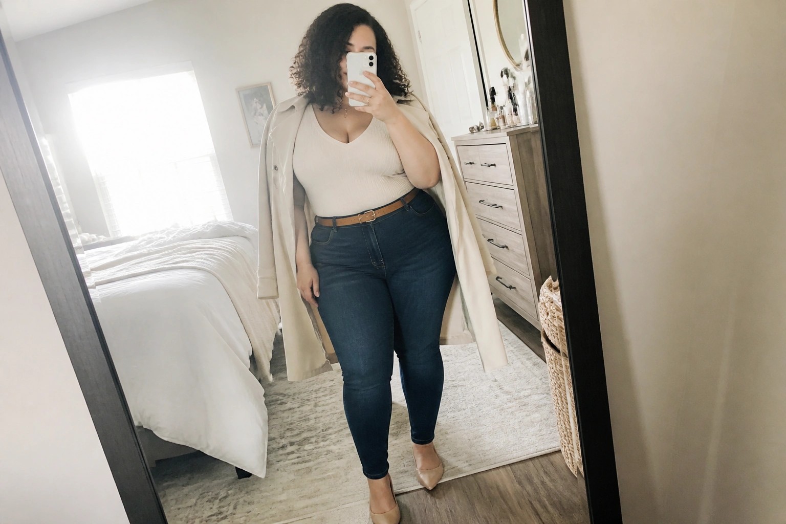 Woman in beige trench coat and jeans taking mirror selfie in bedroom.