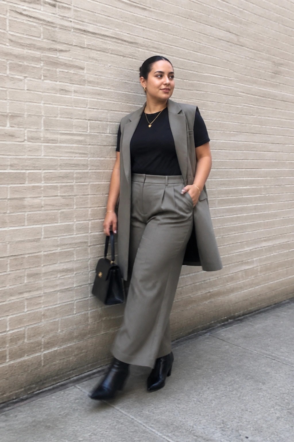 Woman in a long gray sleeveless vest over a black short sleeve top with matching gray wide leg trousers and black ankle boots, holding a small black handbag.