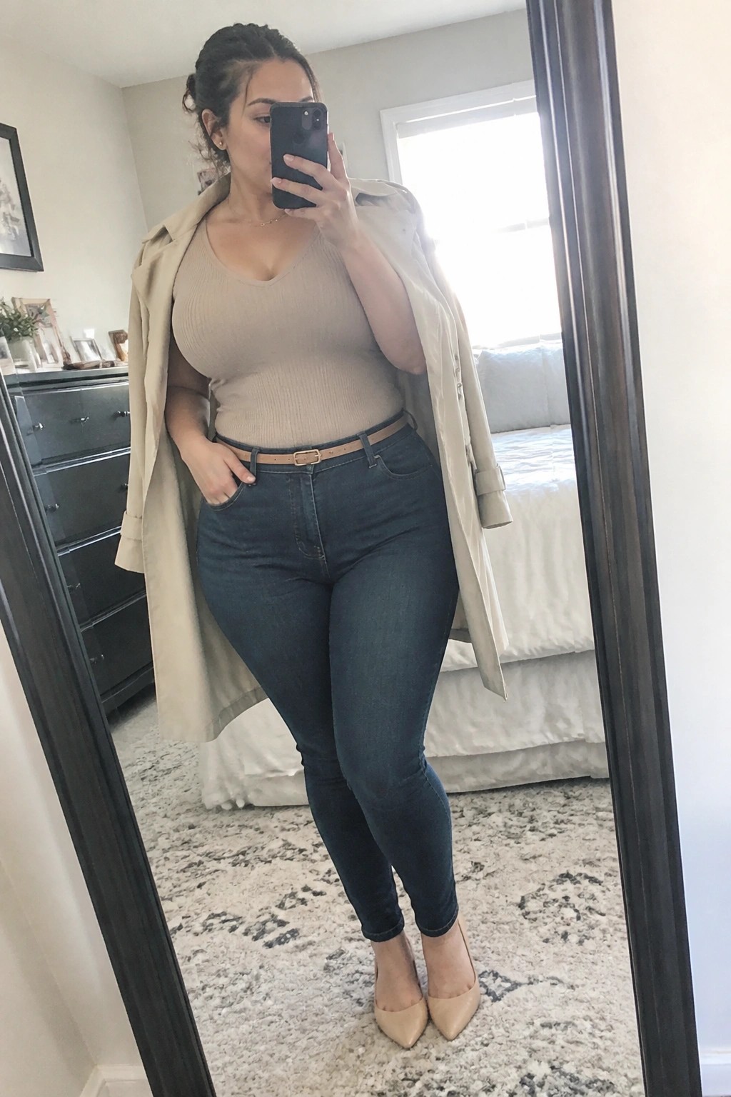 Woman in beige trench coat and jeans taking mirror selfie in bedroom.
