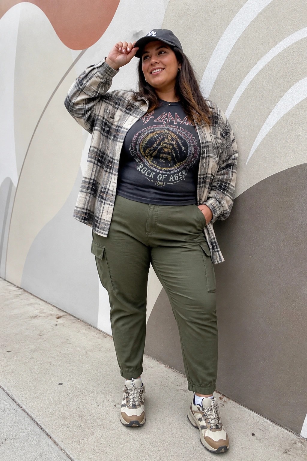 Woman in plaid overshirt, Def Leppard tee, olive cargo pants against mural wall