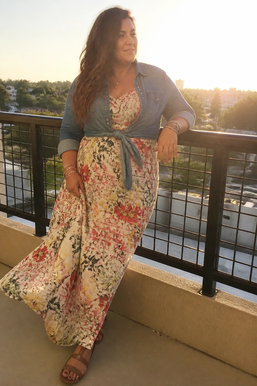 Woman in floral maxi dress with tied denim jacket on sunset balcony