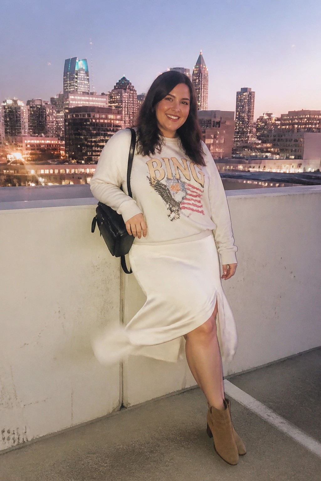 Woman in white sweatshirt and skirt posing on rooftop against dusk city skyline
