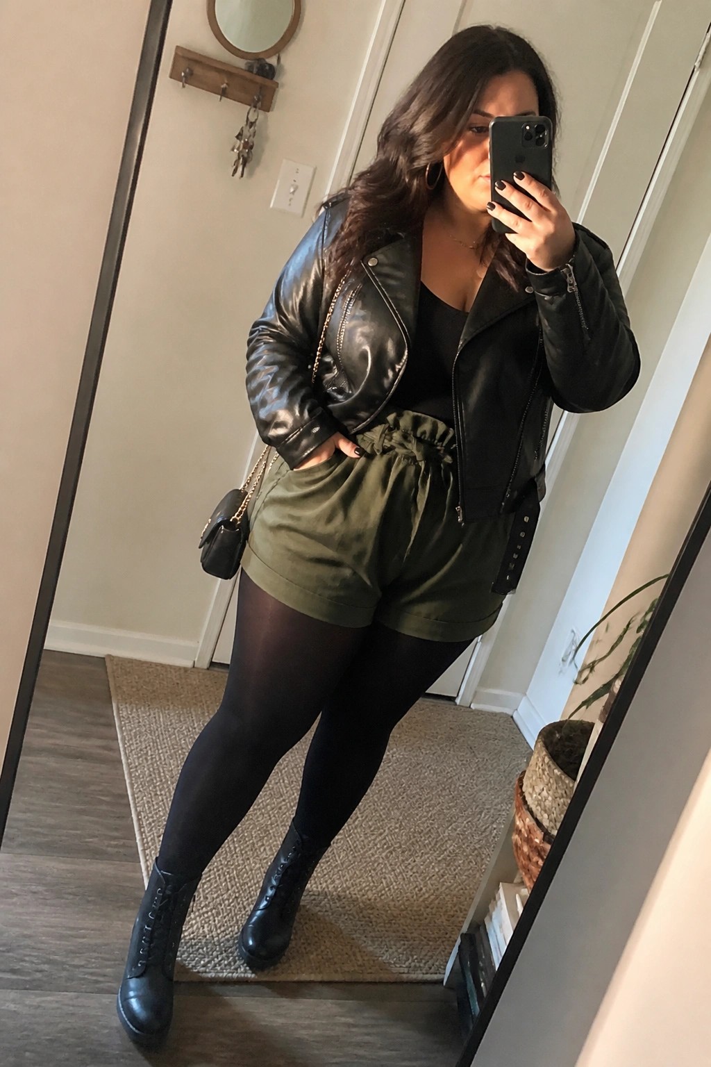 Woman in black leather jacket and olive shorts taking mirror selfie with phone