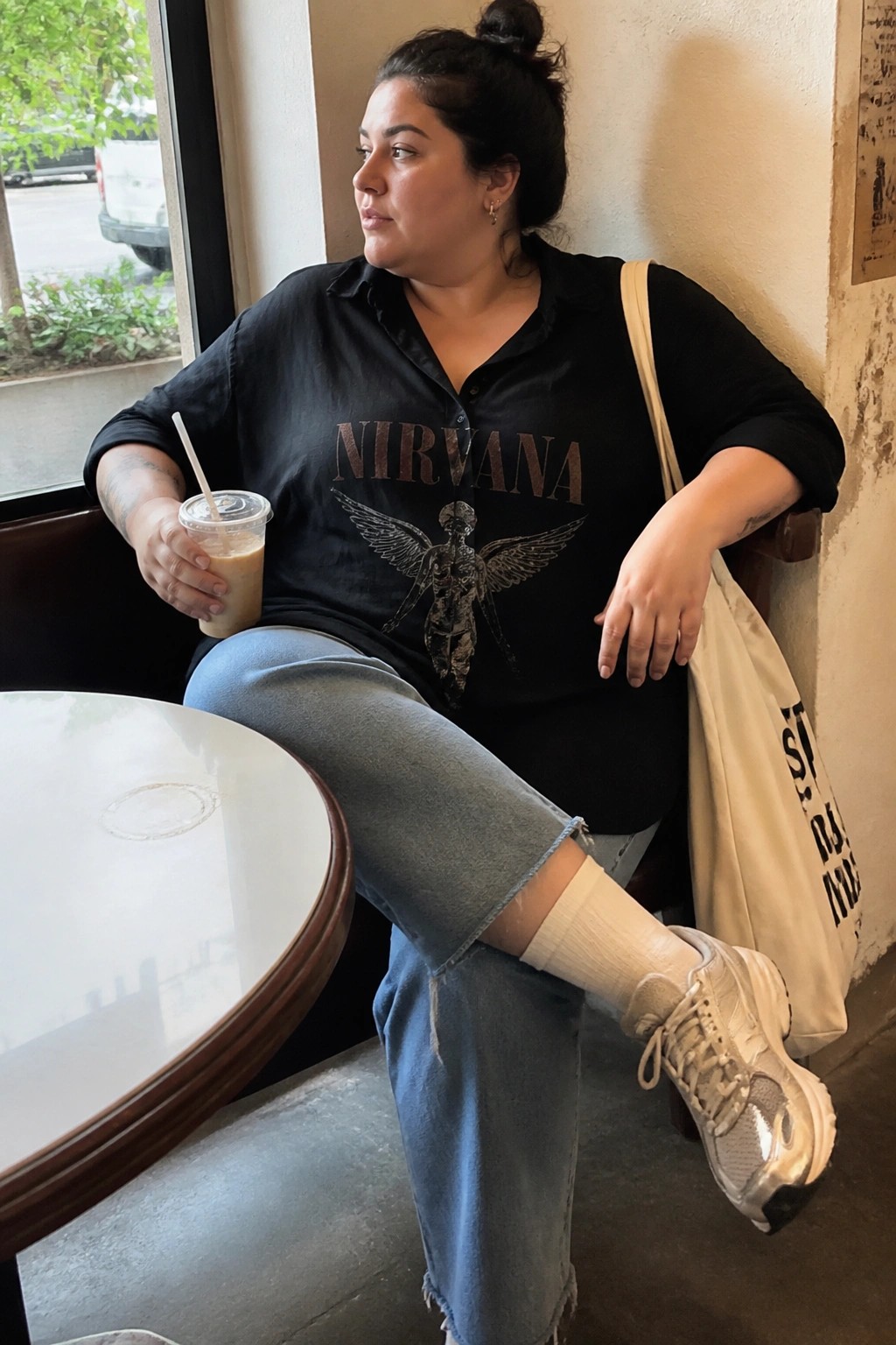 Woman in black Nirvana shirt and jeans holding iced coffee beside canvas tote in cafe