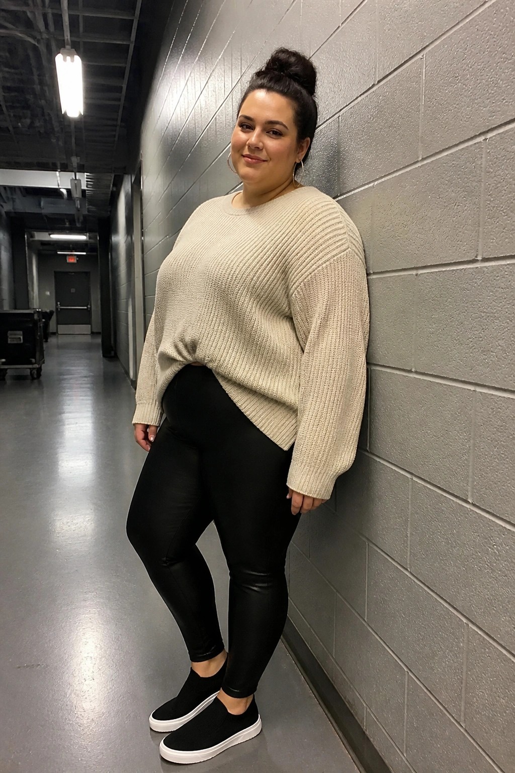 Plus-size woman in beige sweater and black leggings leaning on gray cinderblock wall