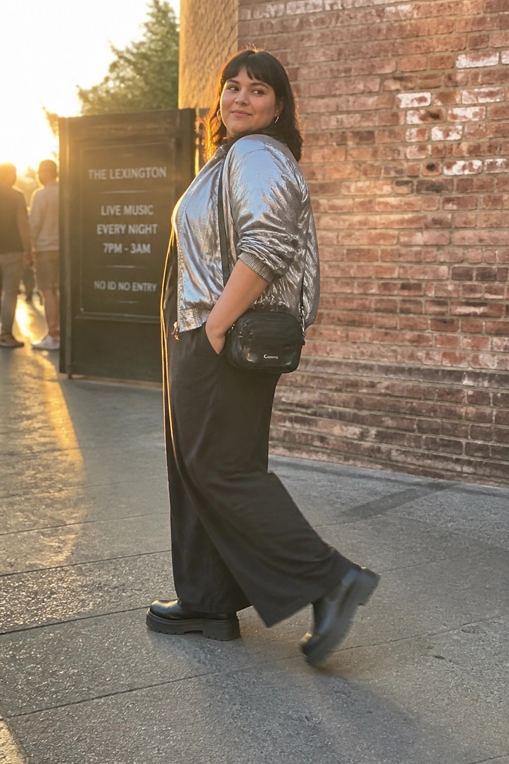 Woman in silver metallic jacket and black wide-leg pants posing by brick wall at sunset