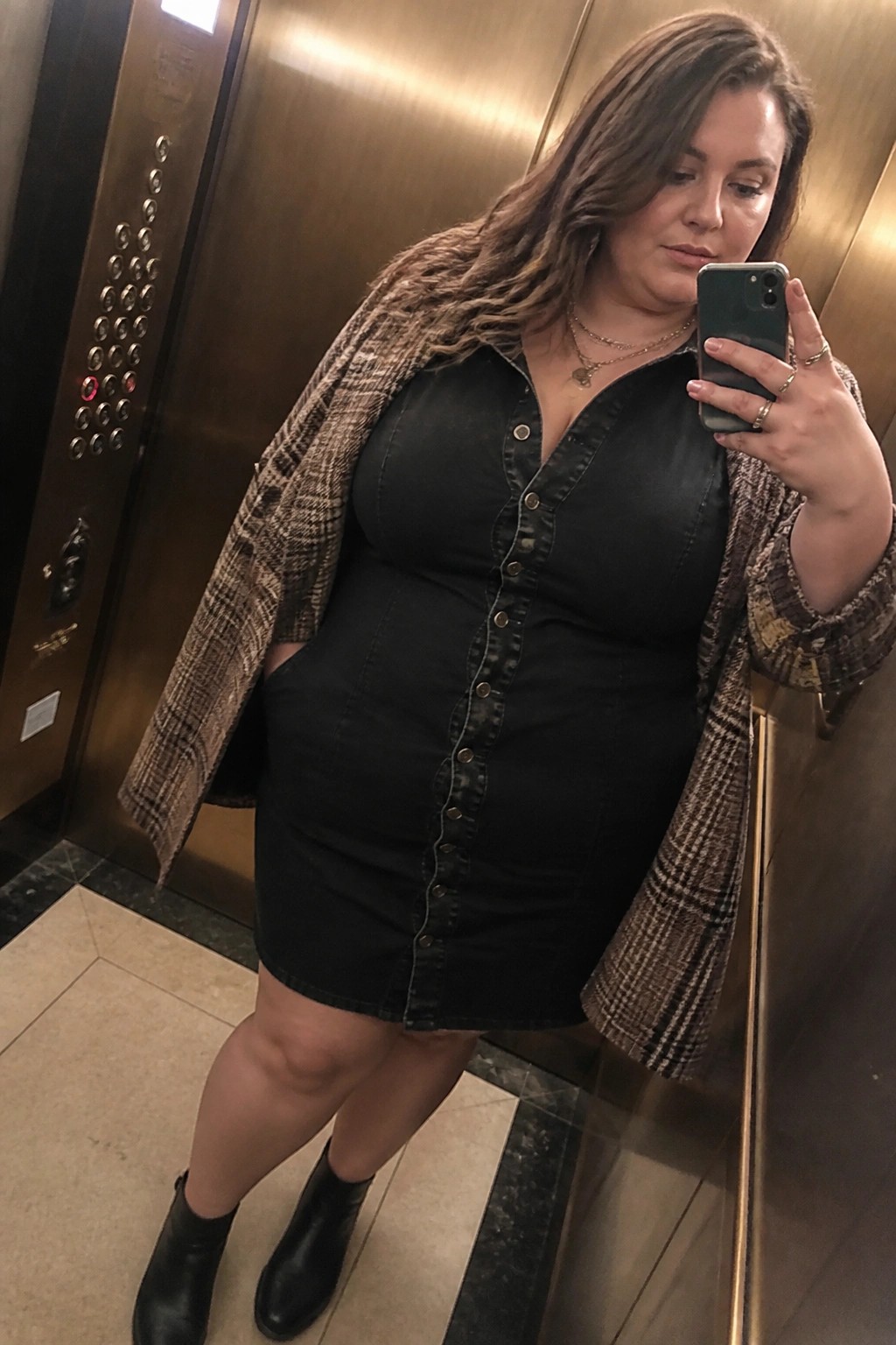 Woman in black denim dress and plaid jacket taking mirror selfie in elevator