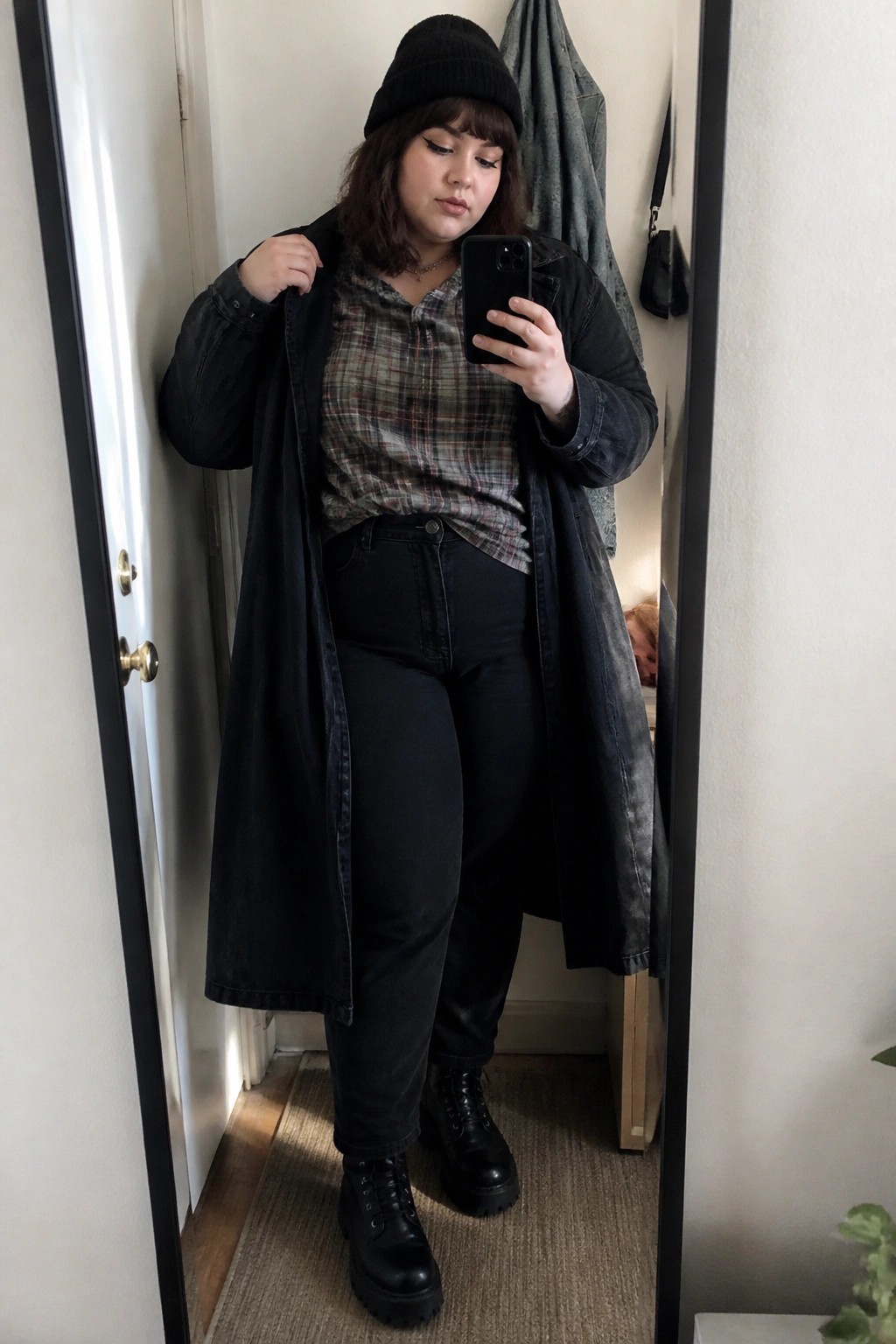 Woman in black beanie, plaid shirt, and long denim coat taking mirror selfie