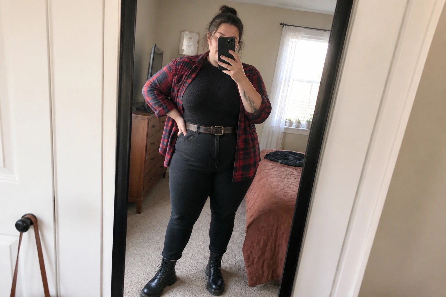 Woman in red-black plaid shirt, black top, jeans, and boots taking mirror selfie