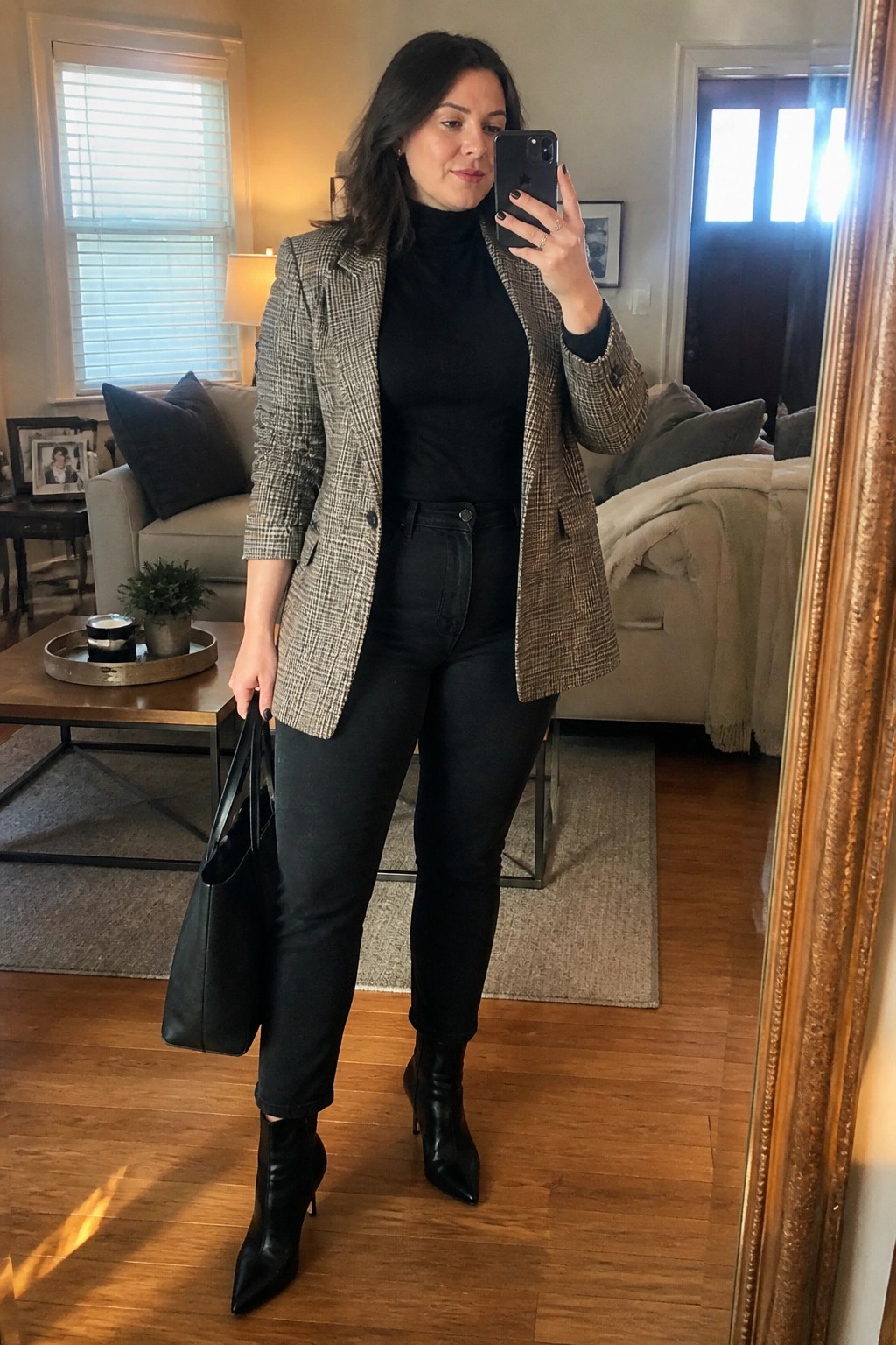 Woman in plus size outfit wearing open plaid blazer over black turtleneck, black denim jeans, and pointed black ankle boots while holding a black tote bag in a mirror selfie.