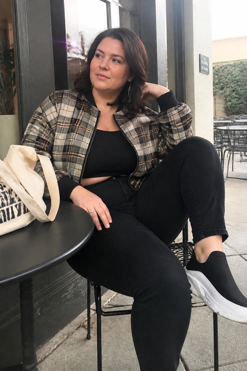 Woman in plaid bomber jacket and black jeans seated at outdoor table with tote bag.