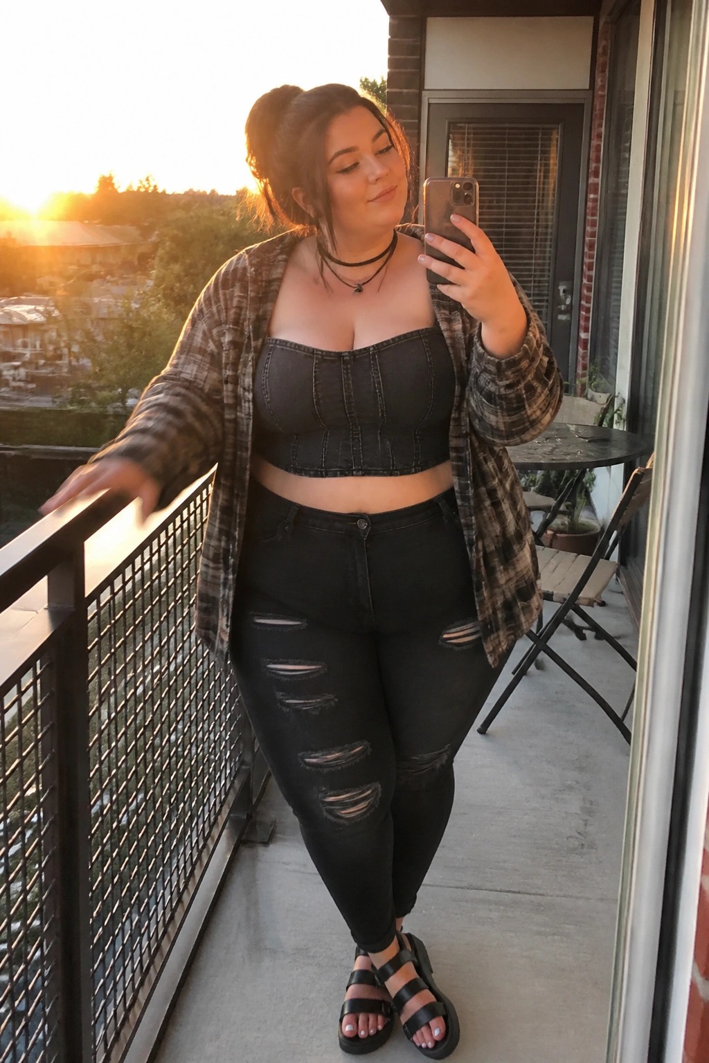 Plus size woman wearing an open brown and black plaid overshirt over a black denim strapless crop top with matching ripped black jeans and black platform sandals while holding a phone for a mirror selfie.