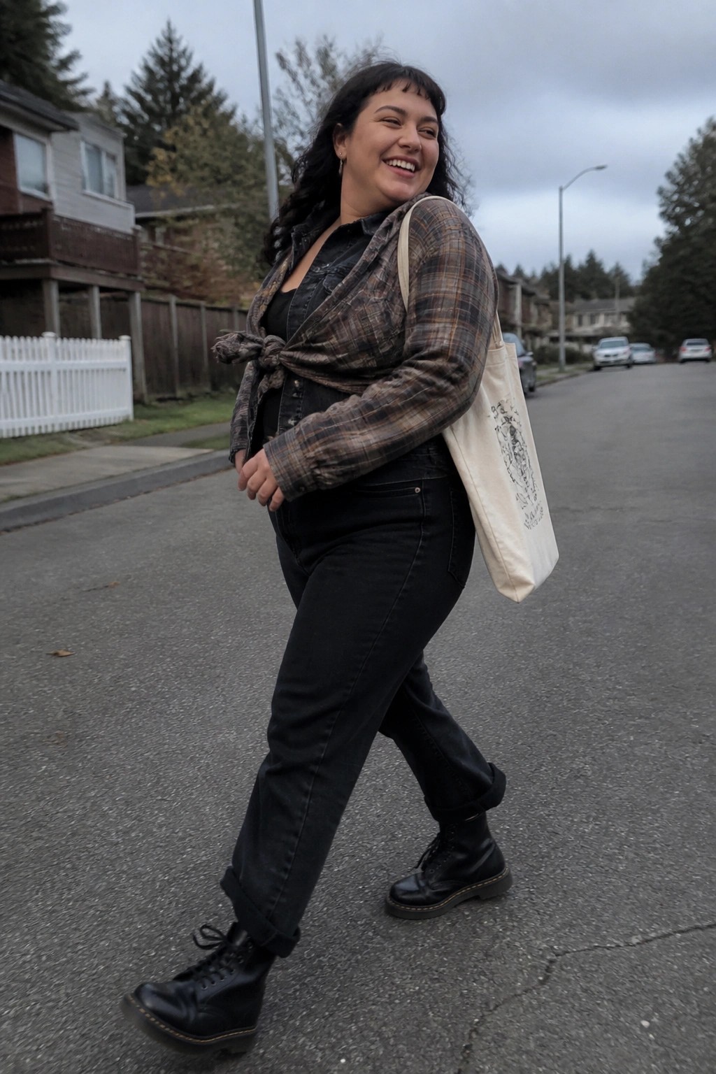 Smiling woman in tied plaid shirt, black jeans, boots carrying canvas tote on street