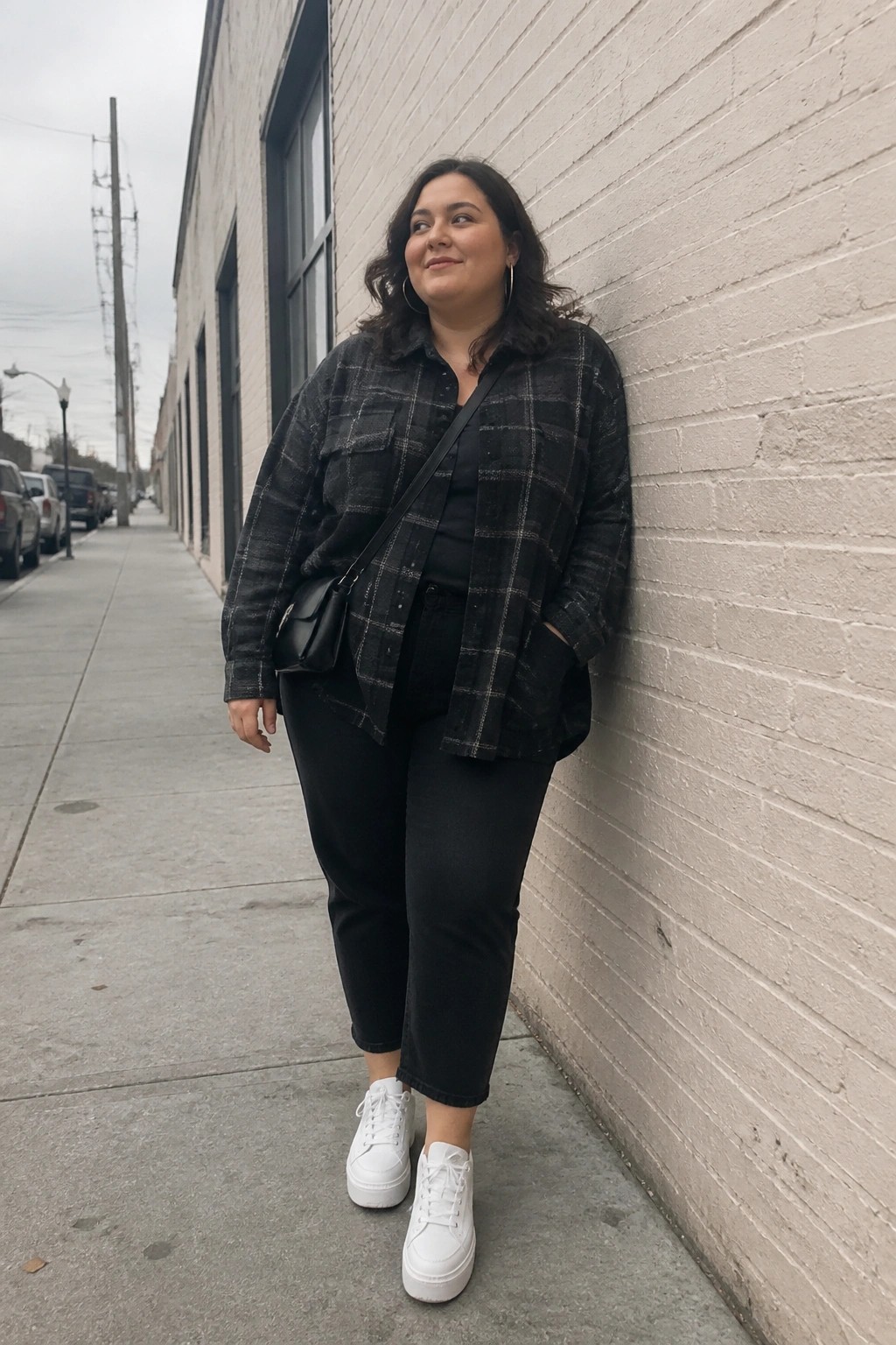 Curvy woman in black plaid shirt leaning against white brick wall on sidewalk.