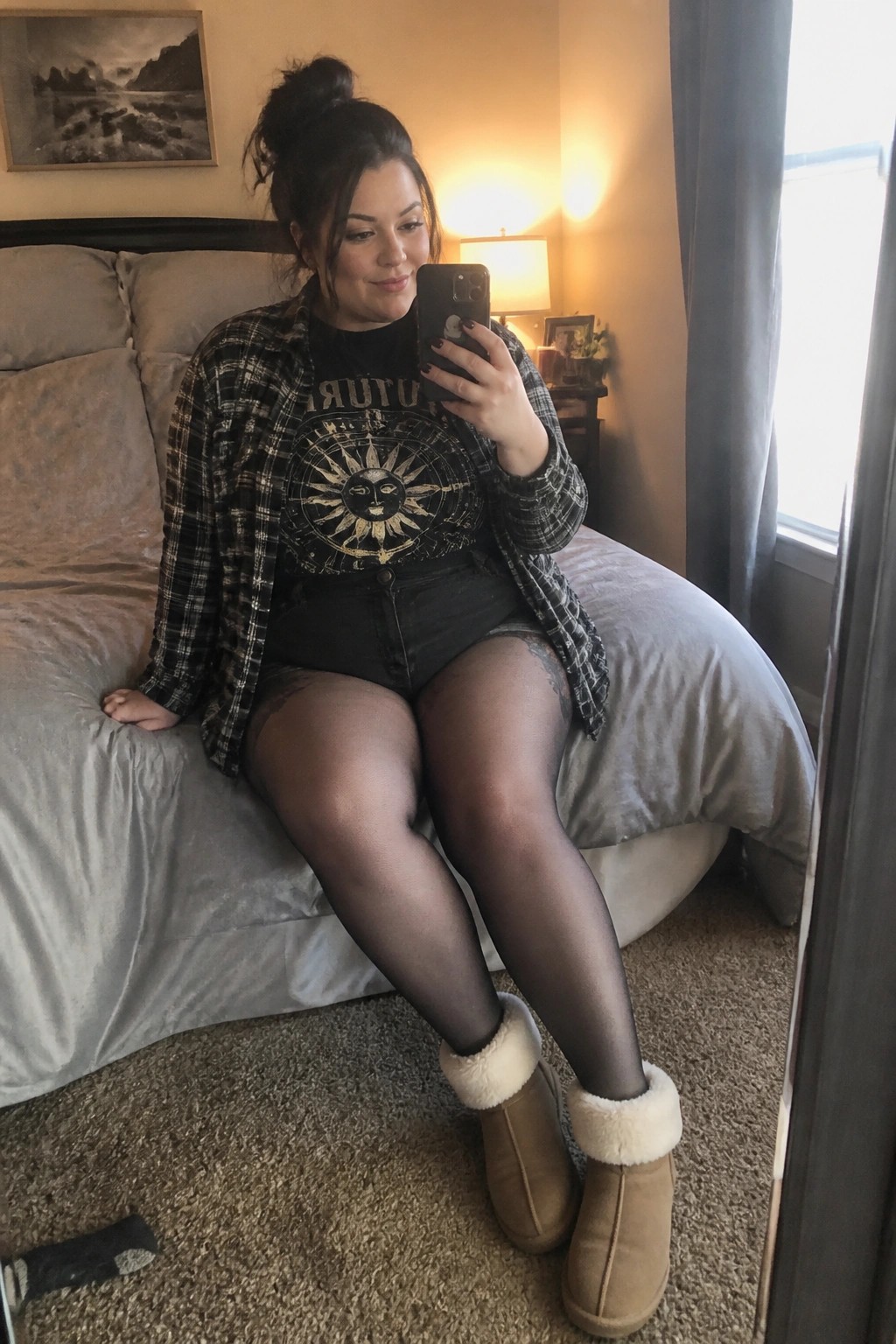 Woman in plaid shirt and boots taking mirror selfie on bed in bedroom