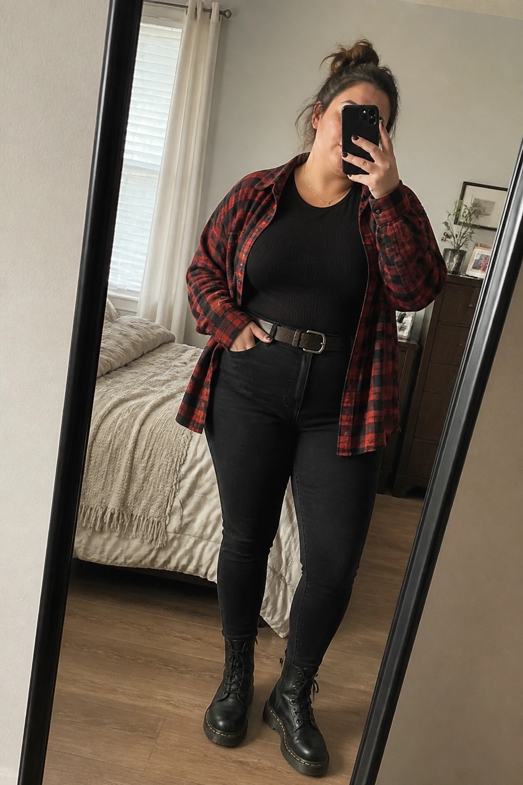 Woman in red-black plaid shirt, black top, jeans, and boots taking mirror selfie