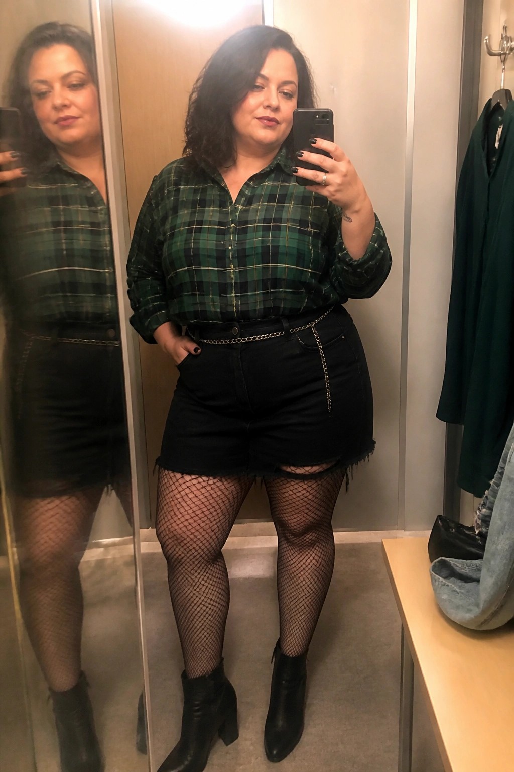 Woman in green plaid shirt and black shorts with fishnets taking mirror selfie.