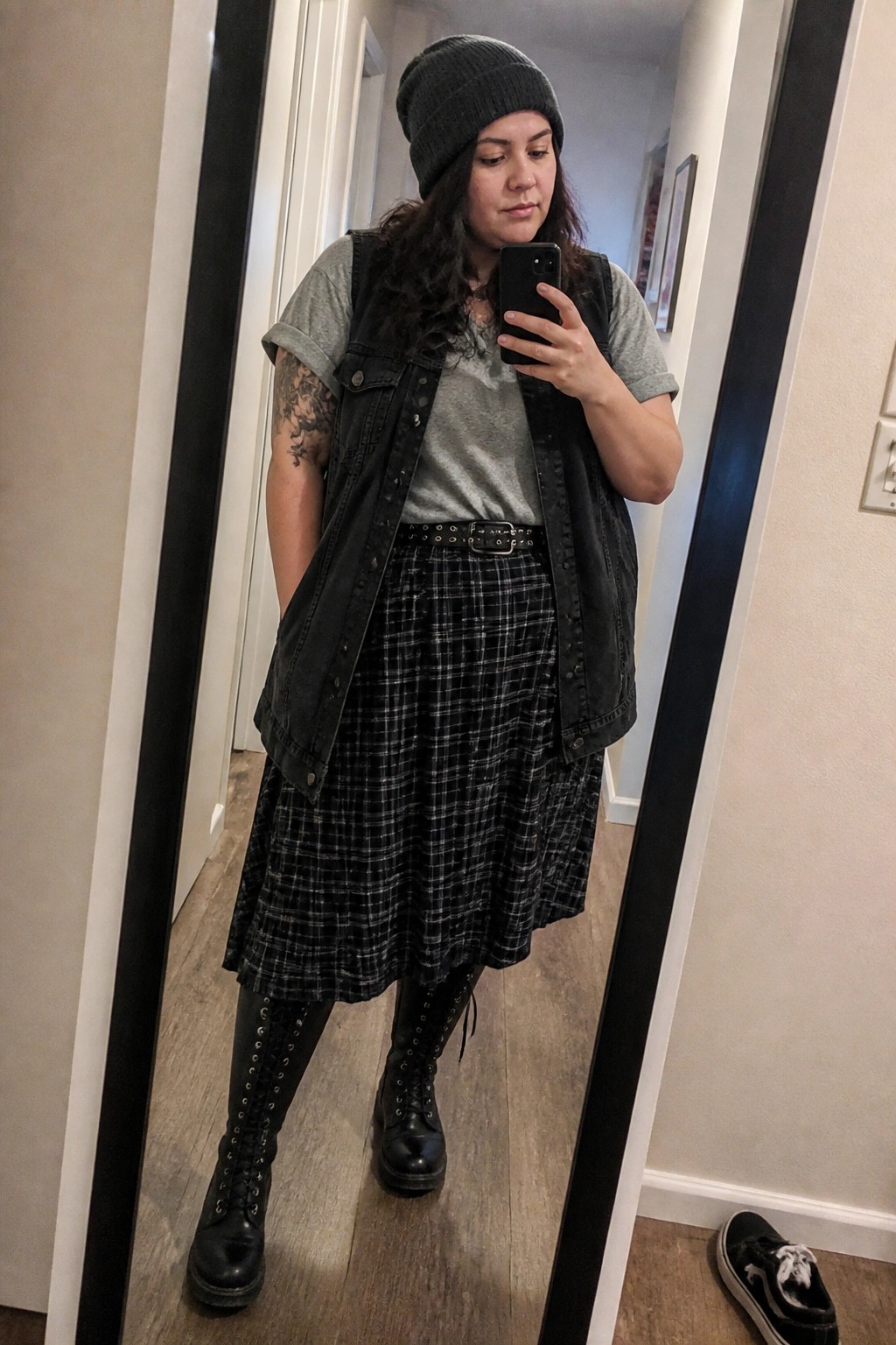 Woman in black beanie, gray tee, denim vest, plaid skirt taking mirror selfie
