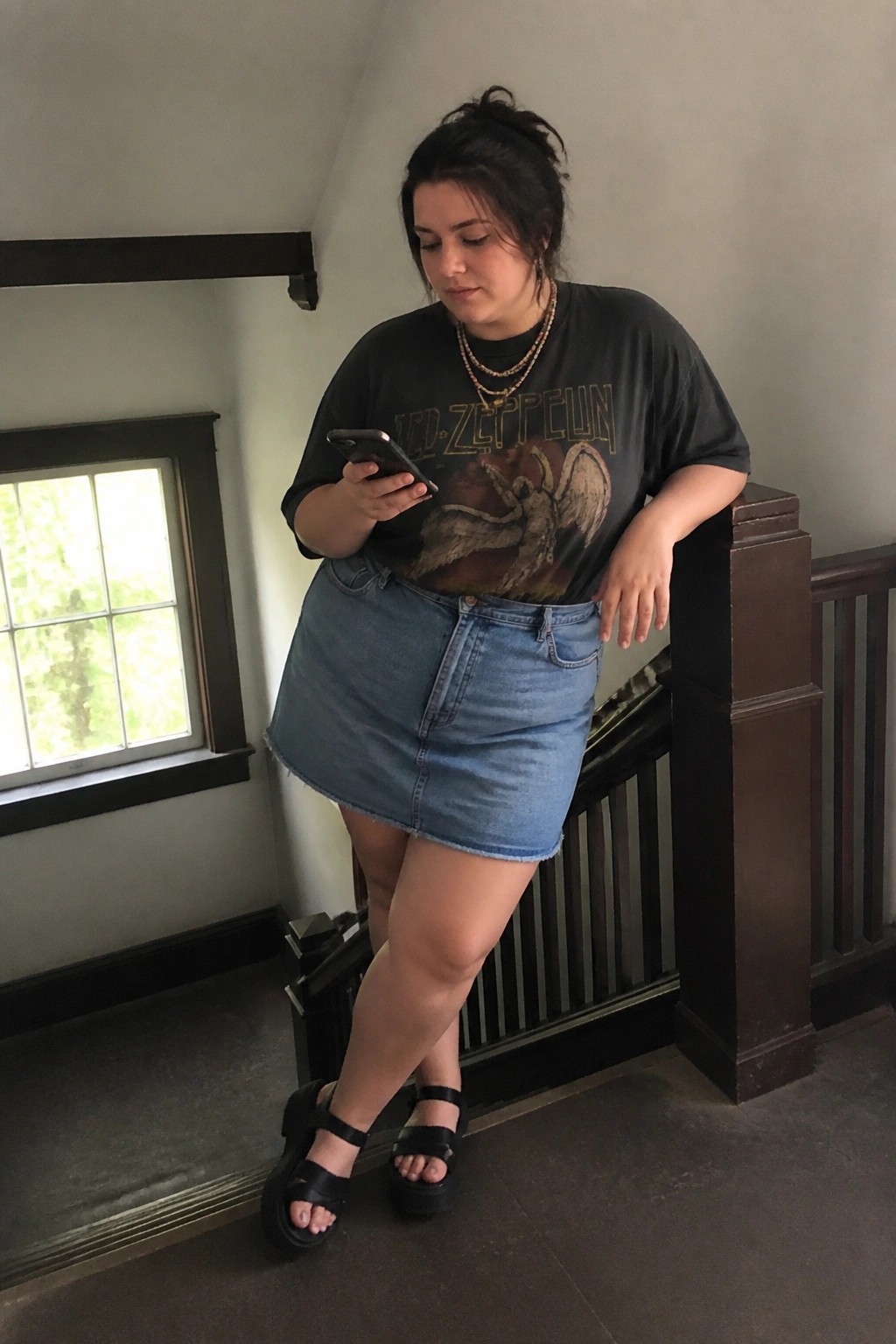 Curvy woman in Led Zeppelin tee and denim skirt checks phone on wooden stairs.
