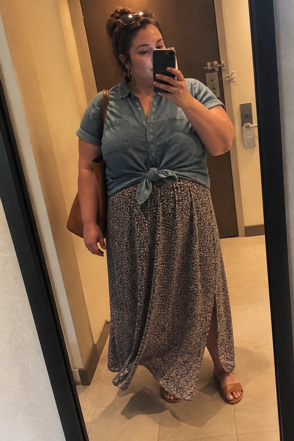 Woman in knotted denim shirt and long patterned skirt taking mirror selfie in hallway