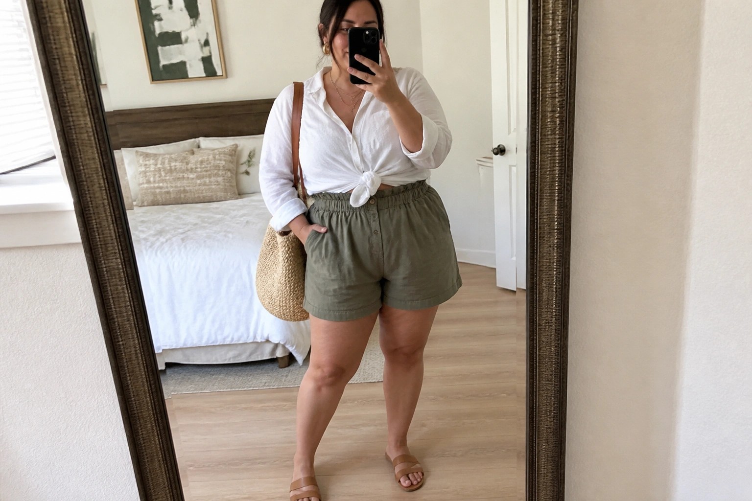 Plus size woman wearing a white button down shirt knotted at the waist with beige tie belt shorts, holding a woven bag and phone for a mirror selfie.