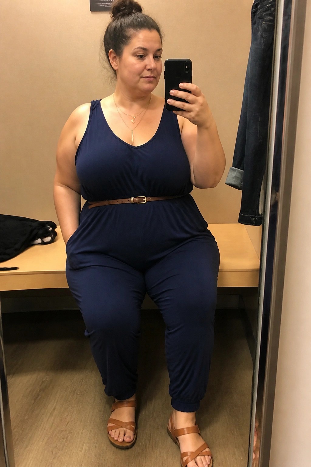 Woman in navy jumpsuit with brown belt taking mirror selfie in fitting room